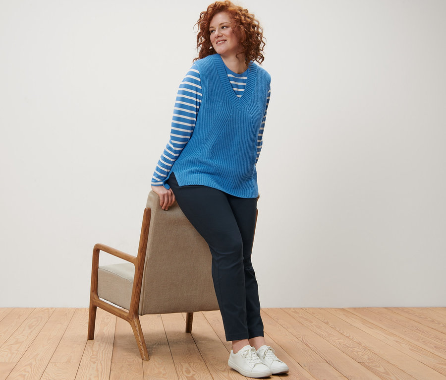 Kıvırcık kızıl saçlı bir kadın sandalyeye yaslanmış, Çizgili Uzun Kollu Tişört, örgü yelek ve koyu mavi pantolon giyiyor.