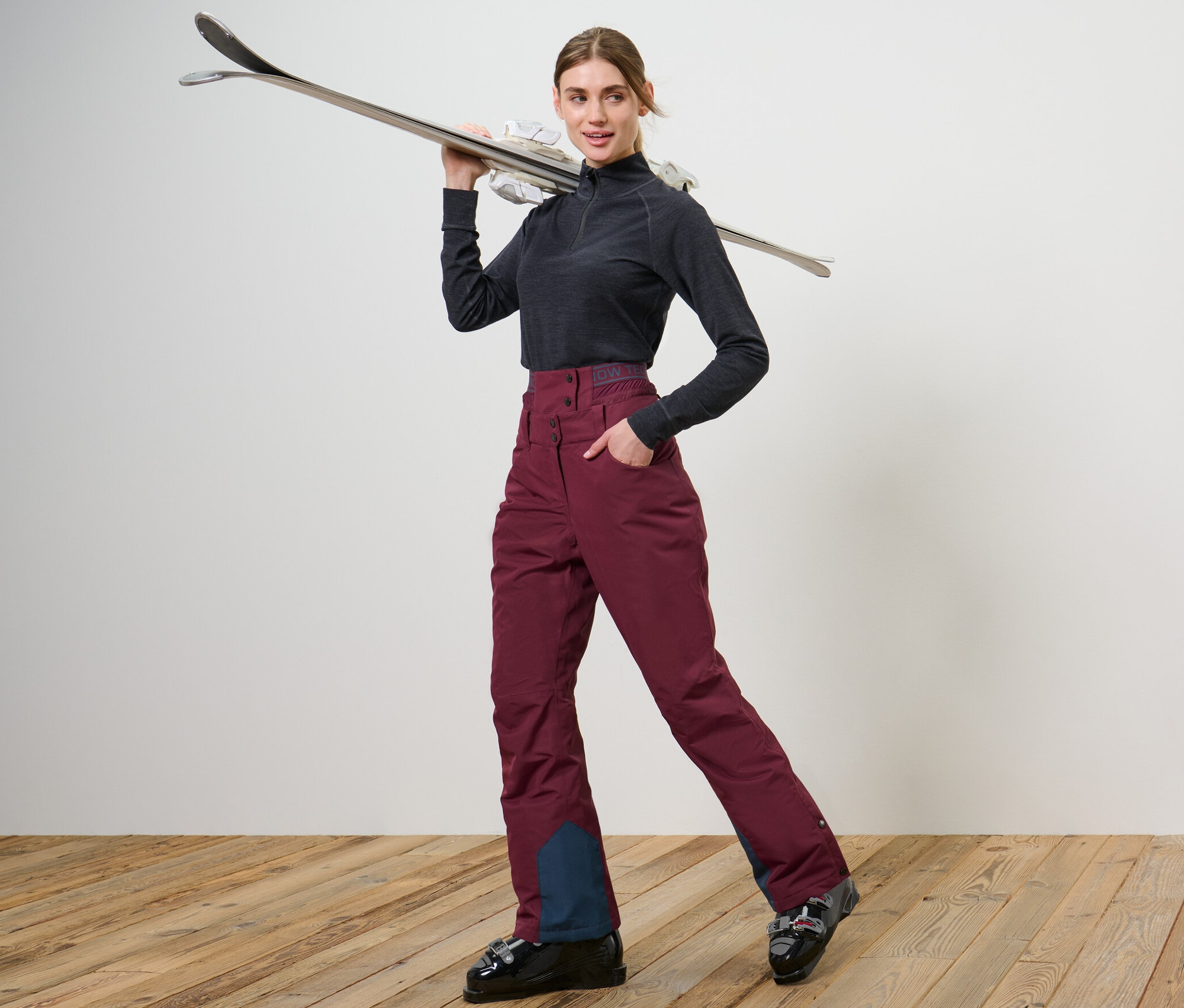 Kadın kayak ayakkabıları ve şarap kırmızısı kayak ve snowboard pantolonuyla poz veriyor.