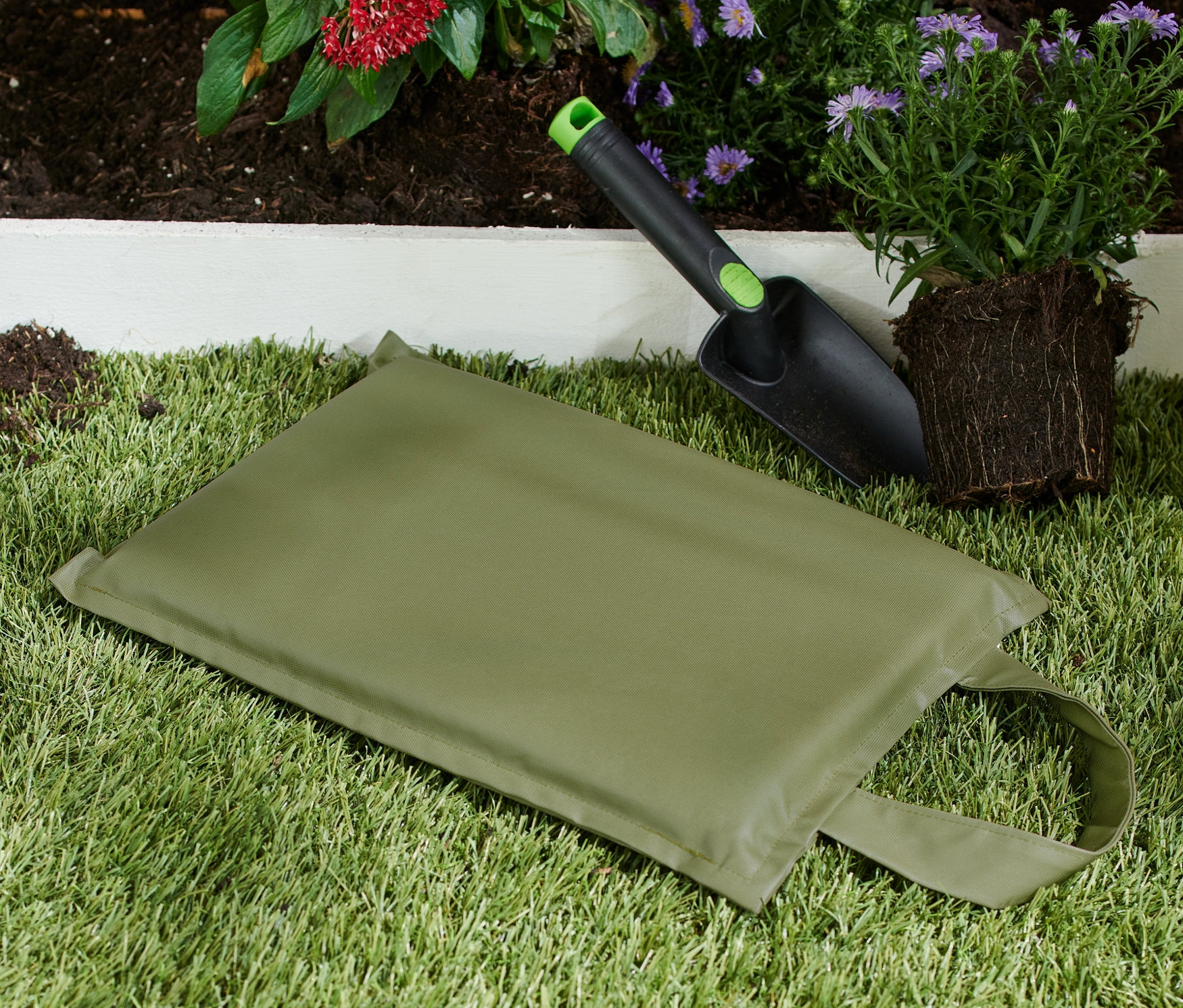 Çim üzerinde bir diz yastığı, kürek ve bitki bulunmaktadır.