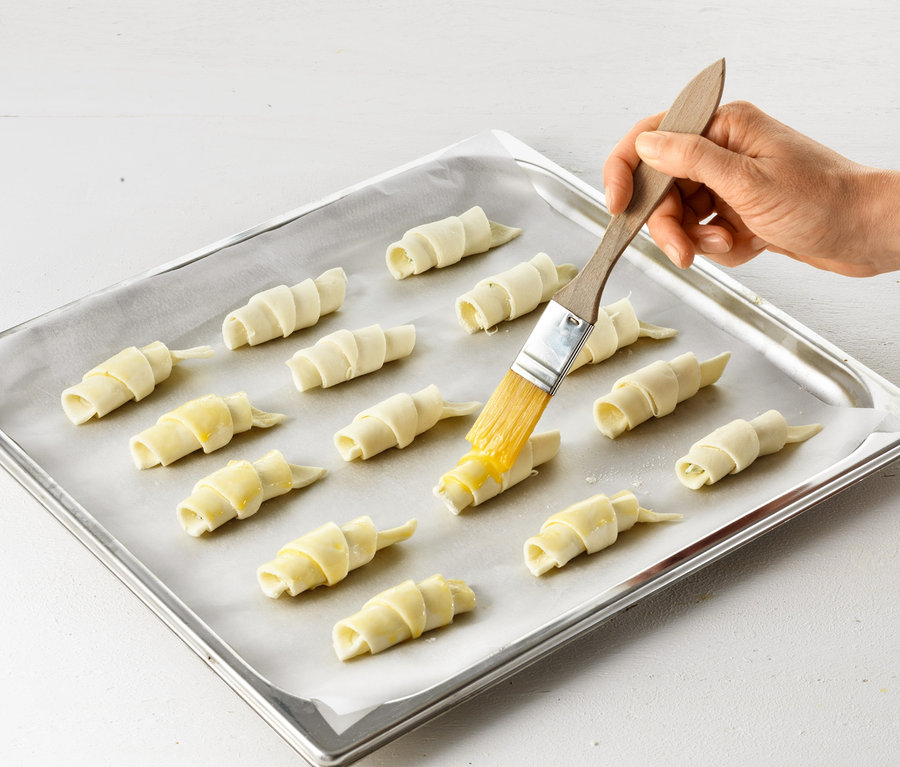 Bir el, fırın tepsisindeki çiğ kruvasanları Kruvasan Rulosu ile yağlıyor.