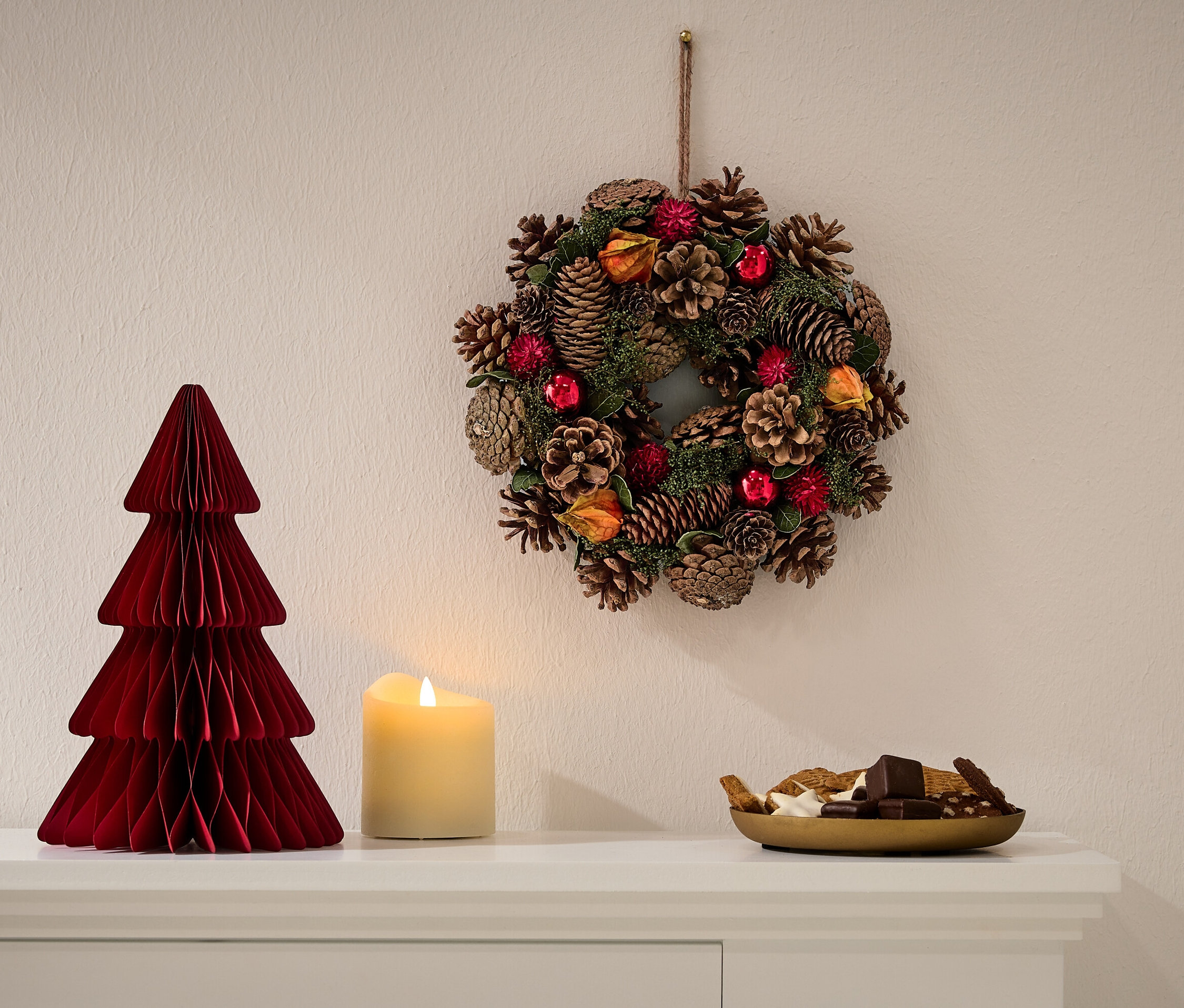 Beyaz bir şifonyerin üzerinde kırmızı bir kağıt yılbaşı ağacı, yanında bir mum ve bir kase şekerleme duruyor. Duvarda bir Noel çelengi asılı.