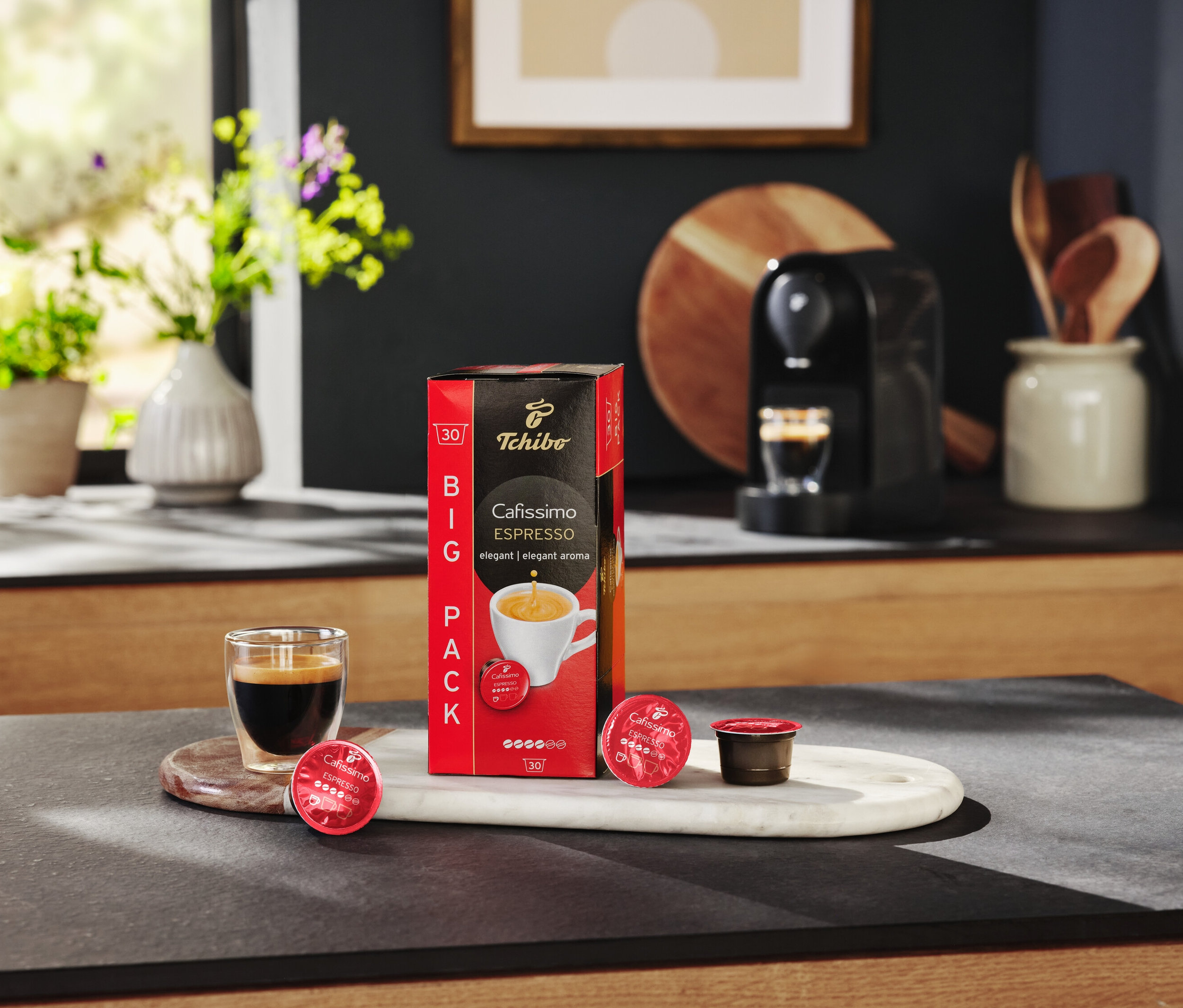 Masanın üzerinde bir paket Cafissimo Espresso kapsülü, bir bardak espresso ve bir kahve makinesi duruyor.