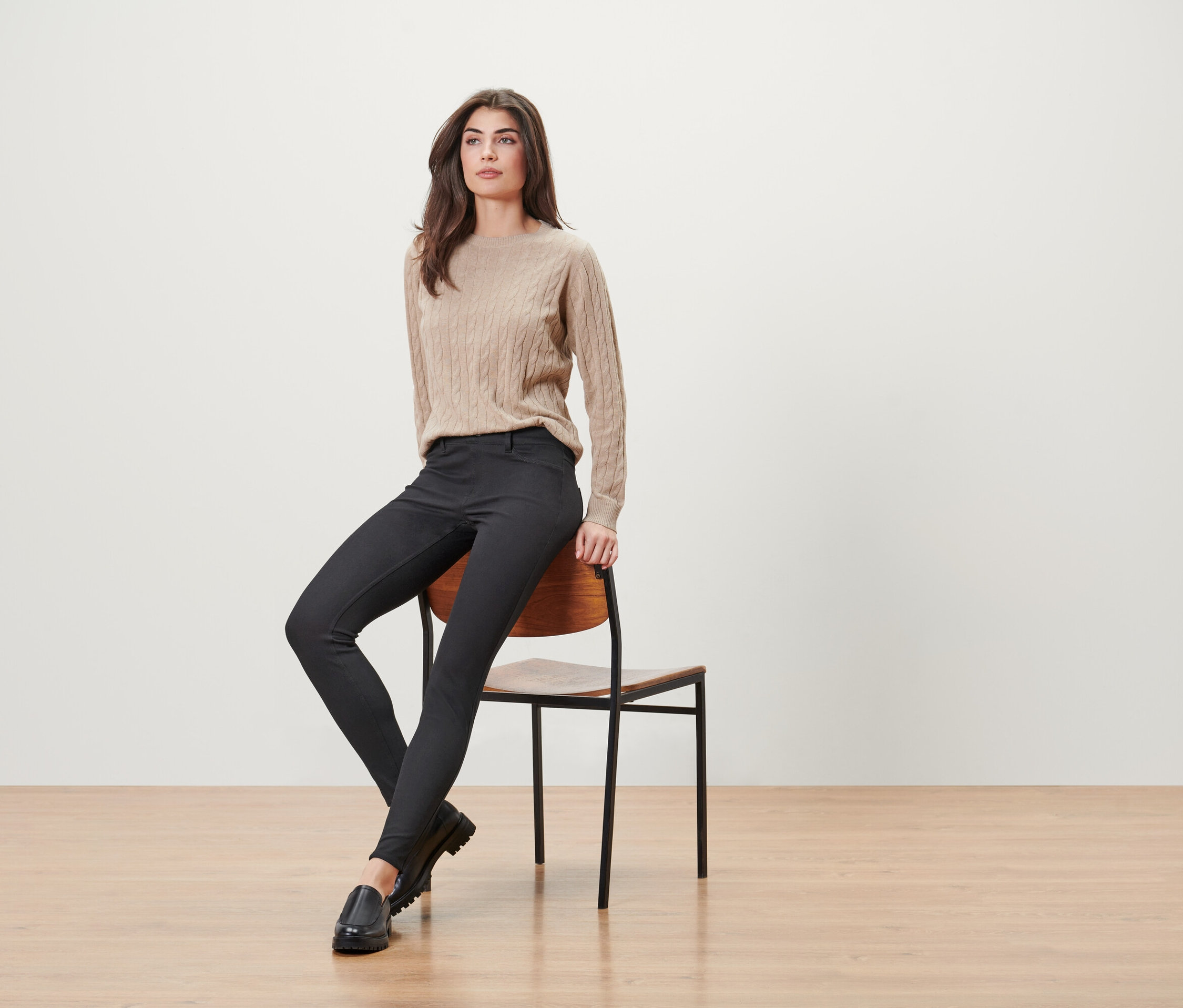 Kadın ahşap bir sandalyede oturuyor, bej bir kazak, siyah pantolon ve siyah mokasen ayakkabılar giyiyor.