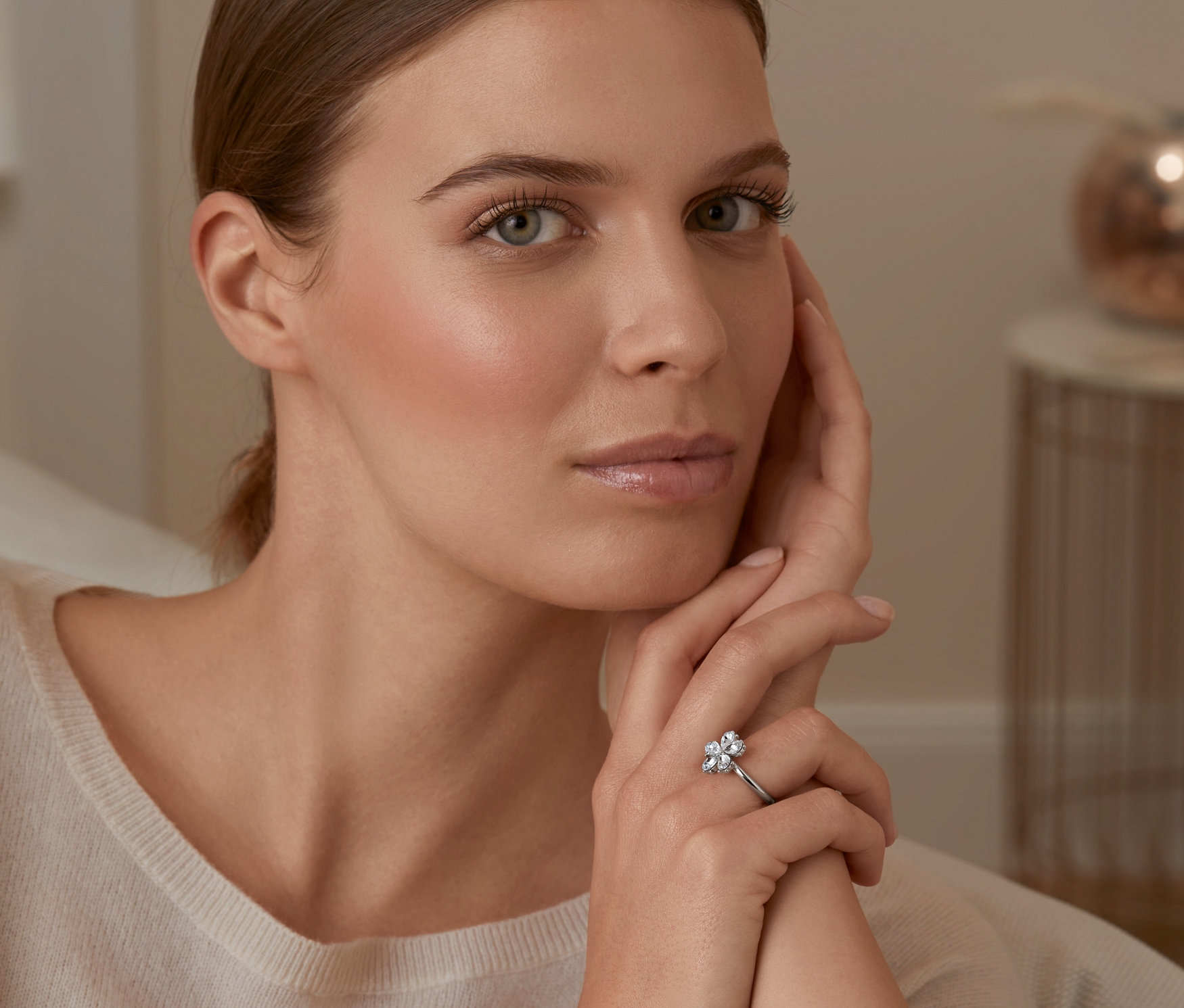 Kadın yüzüne dokunuyor ve parmağında cam kristalden çiçekli rodyum kaplama bir yüzük var.