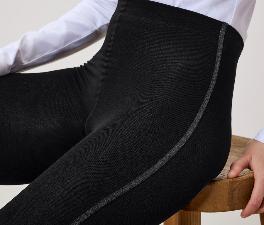 Kadın ahşap bir taburede oturuyor ve yanında gri şerit bulunan siyah bir tayt giyiyor.