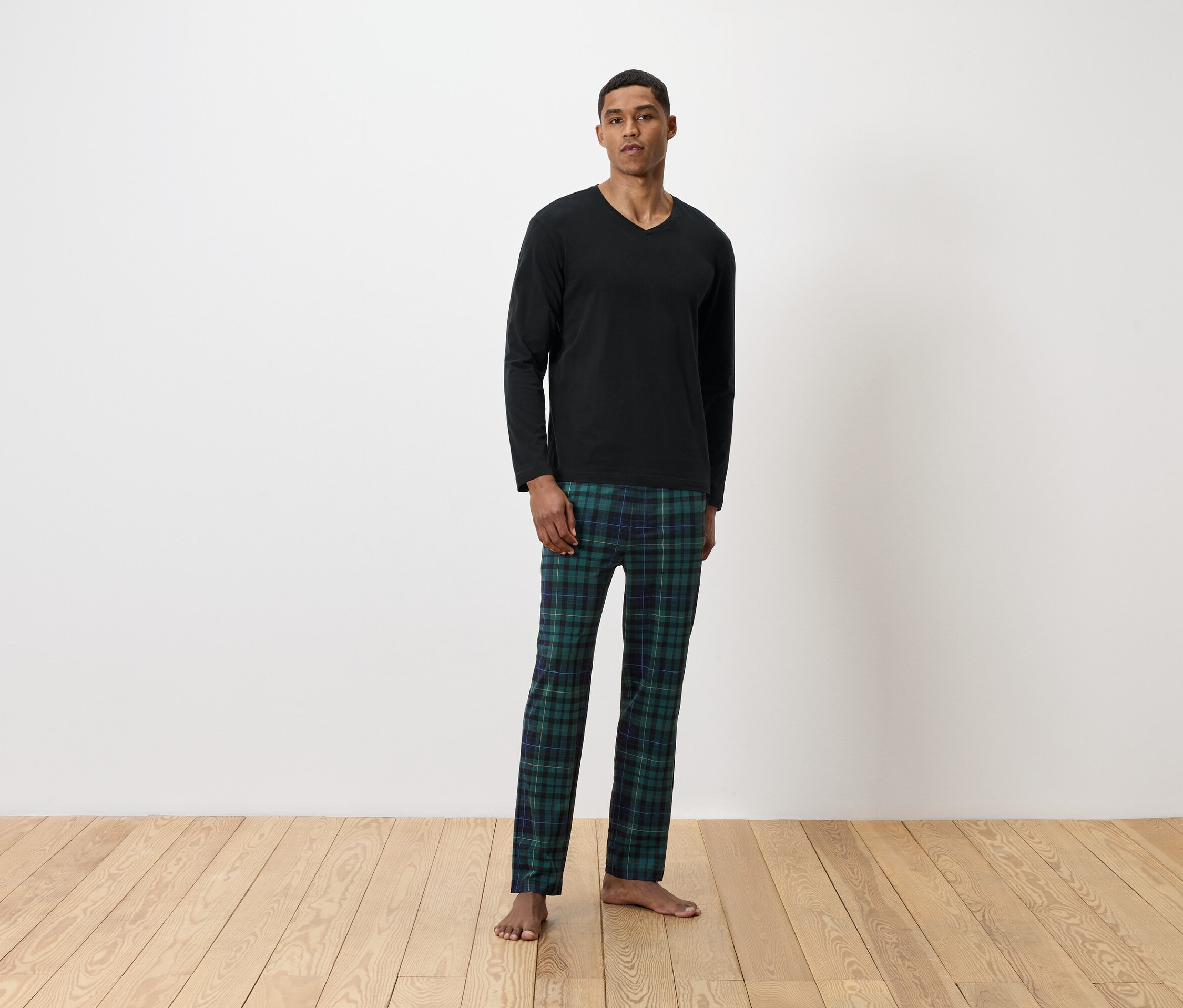 Siyah uzun kollu tişört ve yeşil ekose pijama altı giyen bir adam ayakta duruyor.