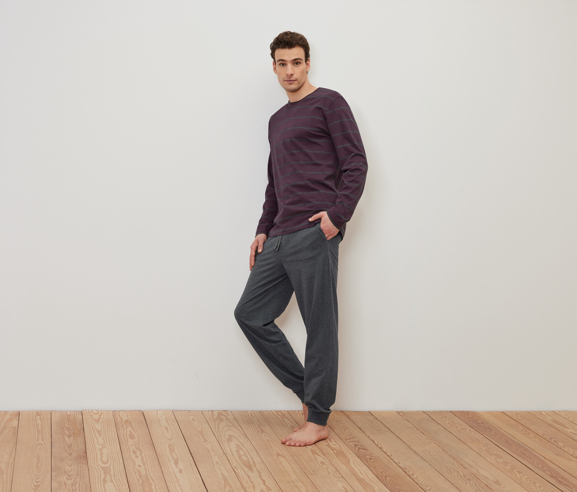 Bir adam ahşap zeminde yalın ayak duruyor, üzerinde mor çizgili uzun kollu bir tişört ve gri eşofman altı var.