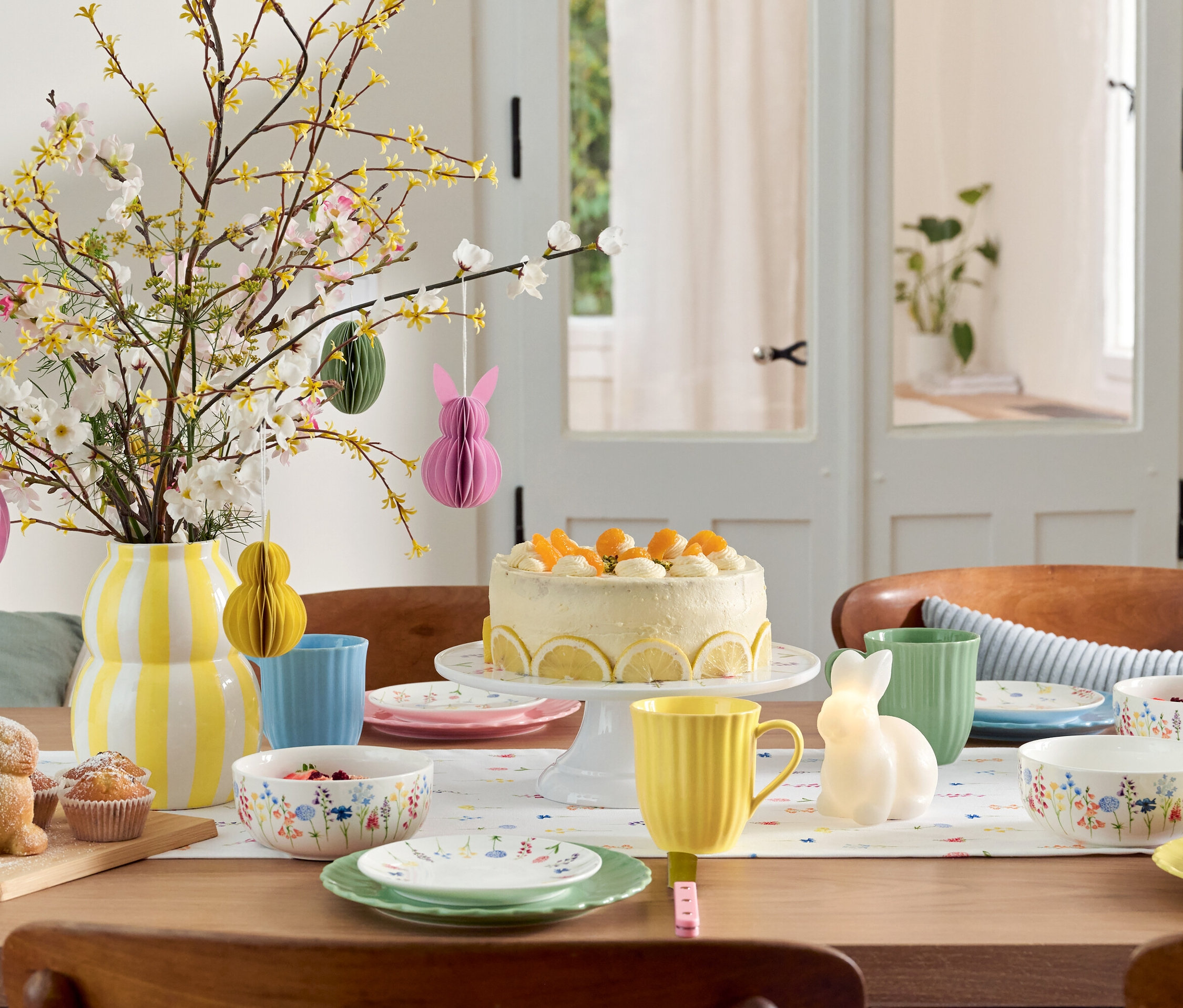 Paskalya dekorasyonları, pasta, sofra takımı ve çiçek vazosu olan yemek masası.