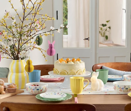 Paskalya dekorasyonları, pasta, sofra takımı ve çiçek vazosu olan yemek masası.