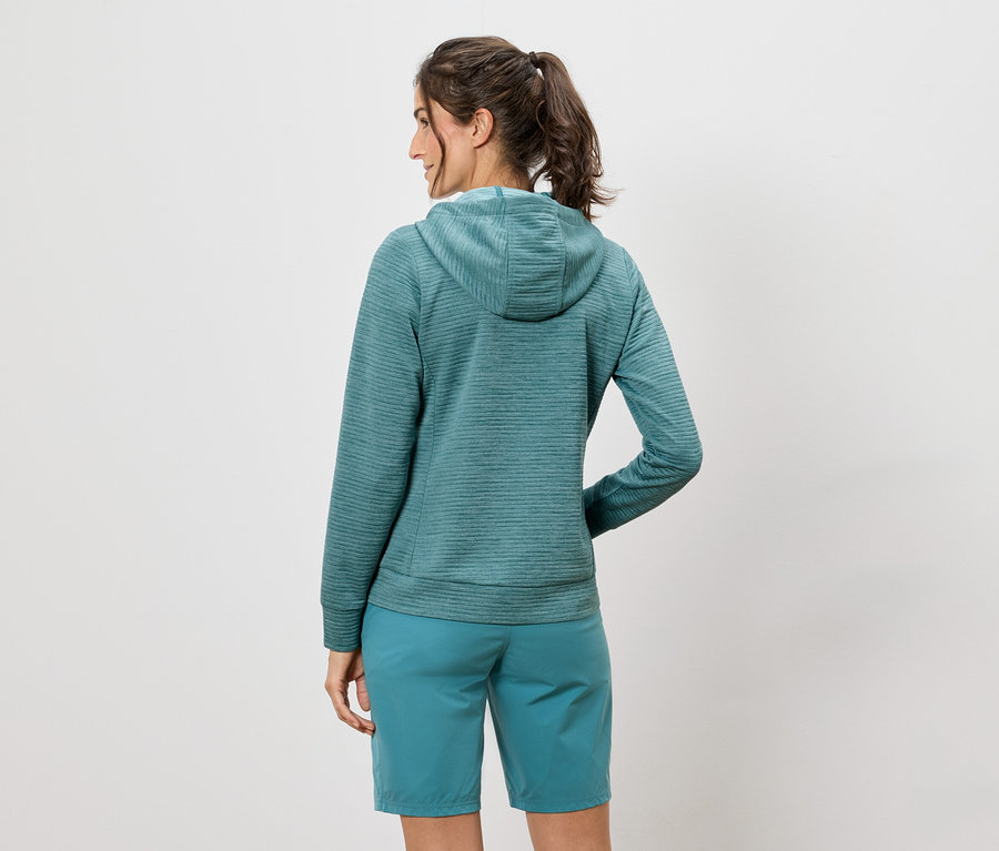 Bir kadın kameraya arkası dönük duruyor, mavi-yeşil kapüşonlu bir sweatshirt ve şort giyiyor.