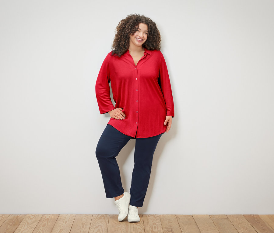 Kıvırcık saçlı kadın kırmızı bluz, mavi pantolon ve beyaz spor ayakkabı giyiyor, beyaz bir duvarın önünde poz veriyor.