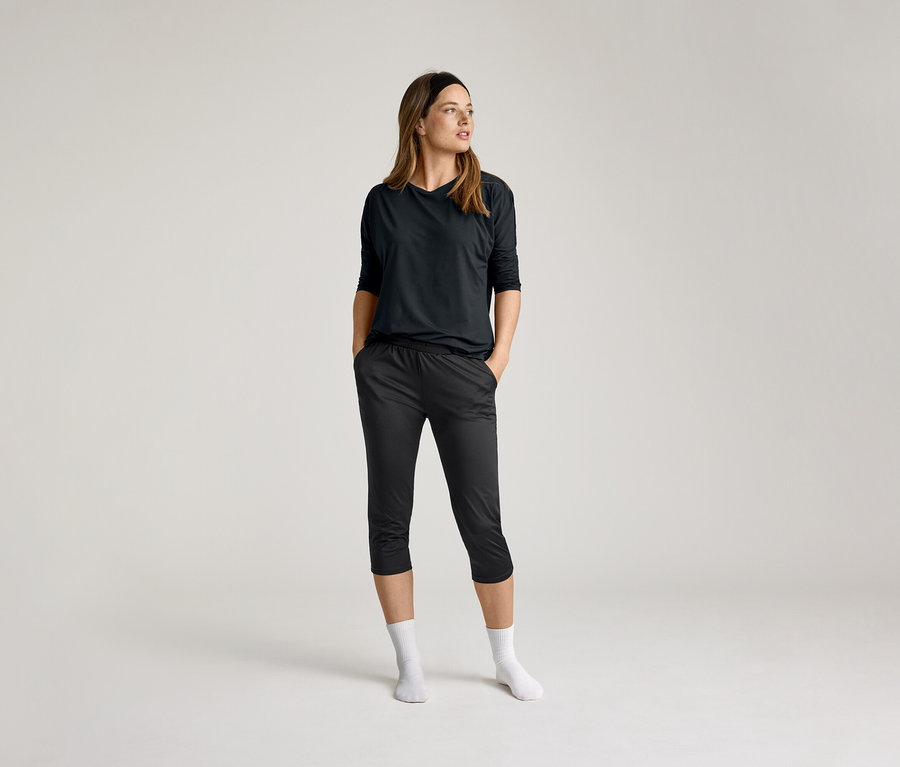 Açık renkli bir arka plan önünde, elleri cebinde, siyah 3/4 spor tişörtü ve koyu renkli pantolon giyen, beyaz çoraplı kadın.