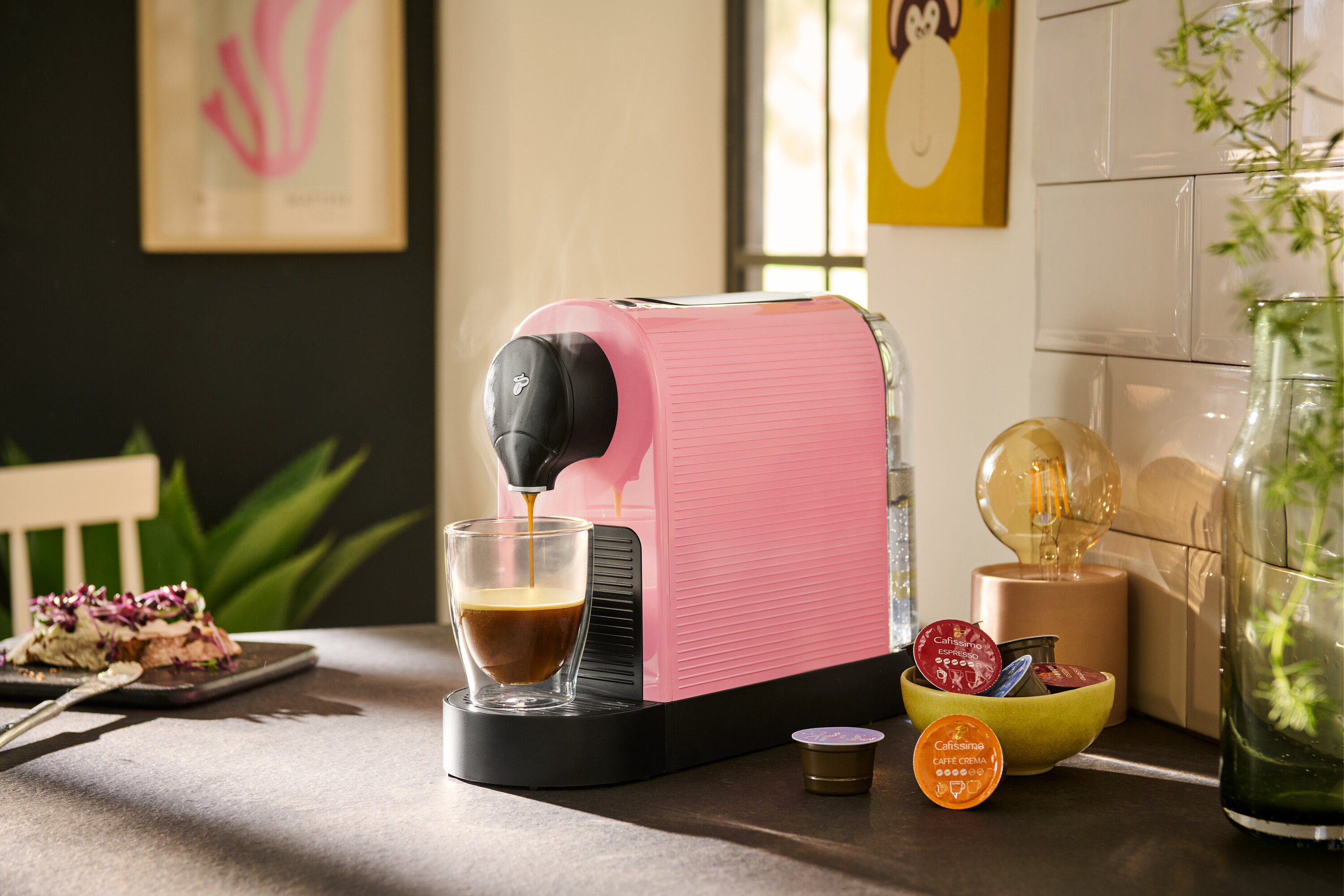 Kiraz çiçeği rengindeki Cafissimo pure+ kahve makinesi bir bardağa kahve koyuyor. Yanında bir kasede kapsüller bulunuyor.