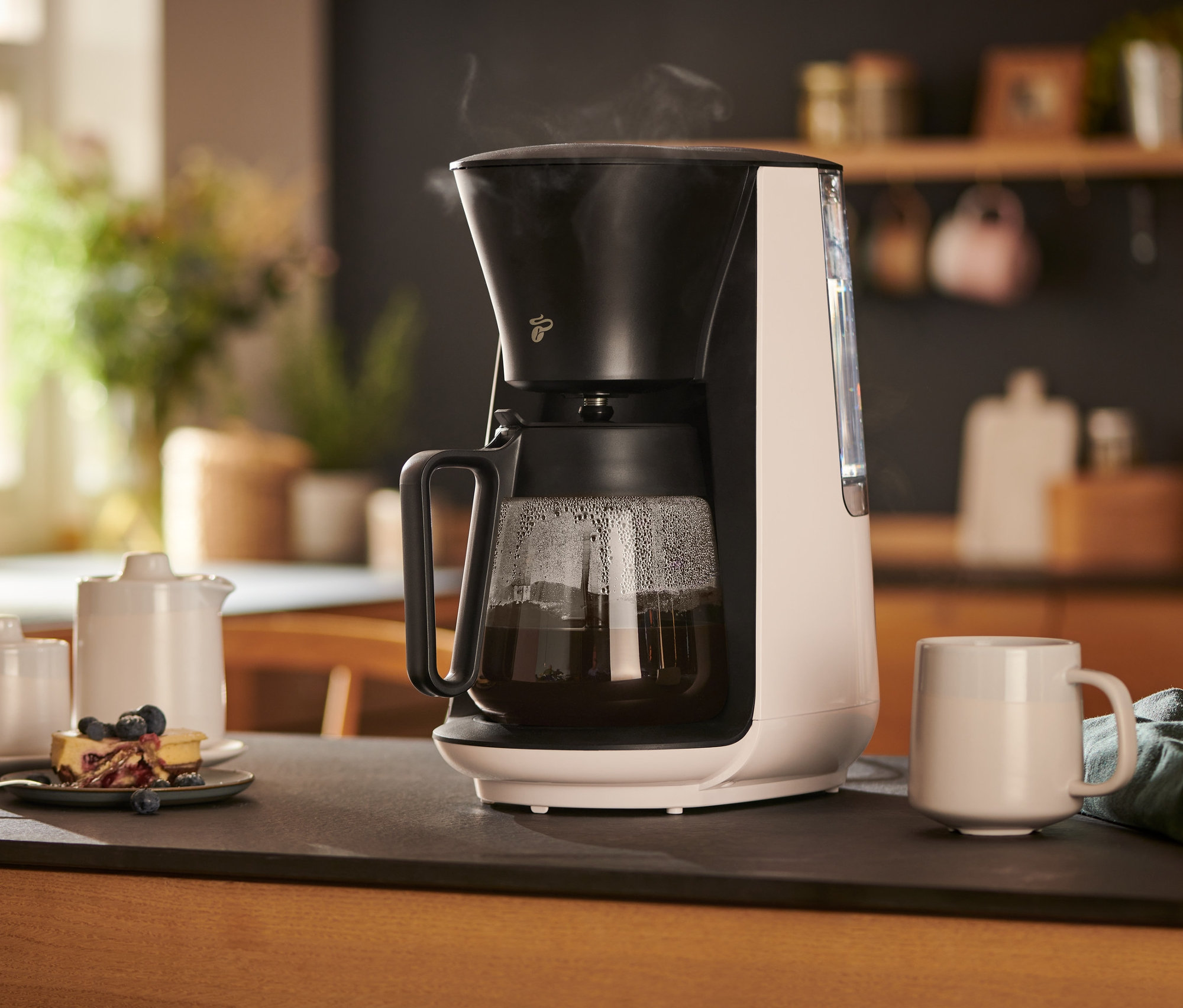 Beyaz »Let's Brew« Filtre Kahve Makinesi bir fincan, sütlük, şekerlik ve bir tabak kekin yanında bir masanın üzerinde duruyor.