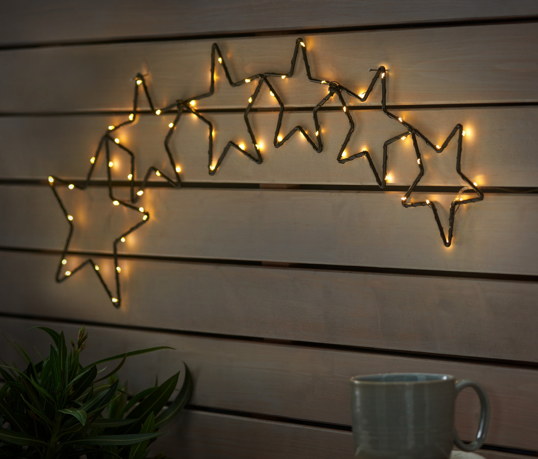 Ahşap bir duvarda LED Outdoor Yıldız Silüeti asılı duruyor. Ahşap bir masanın üzerinde bir fincan ve yıldız şeklinde kurabiyeler bulunan bir tabak var.