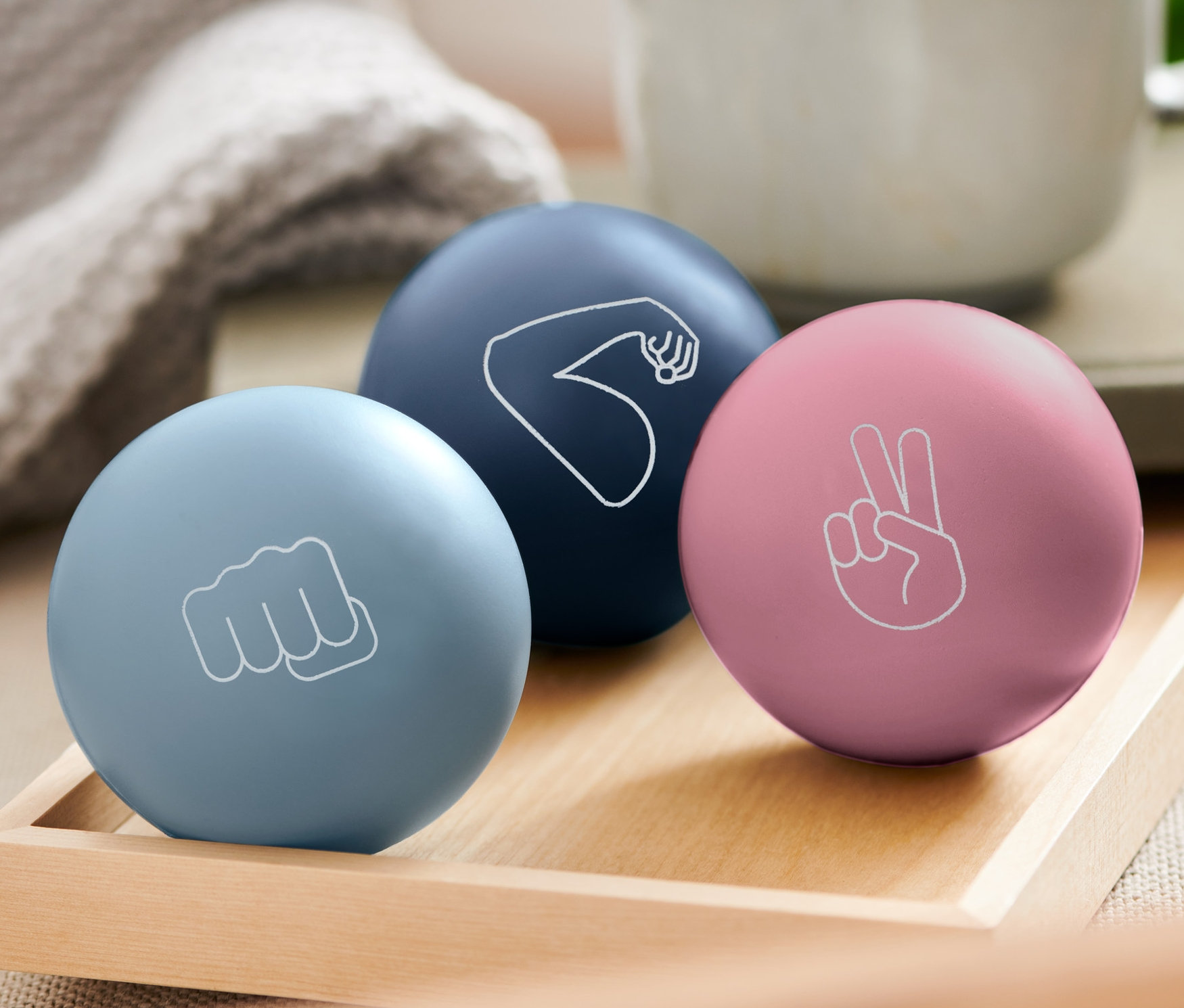 Ahşap bir tepside üç adet stres topu duruyor: açık mavi olanın üzerinde yumruk, koyu mavi olanın üzerinde kol ve pembe olanın üzerinde barış işareti var. Arka planda bir kupa bulunuyor.