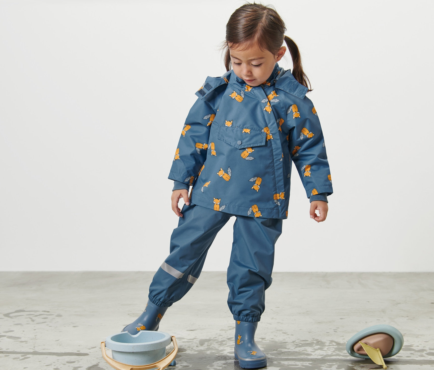 Tilki baskılı çocuk yağmurluğu giyen kız çocuğu bir kova ile su birikintisinin yanında duruyor.