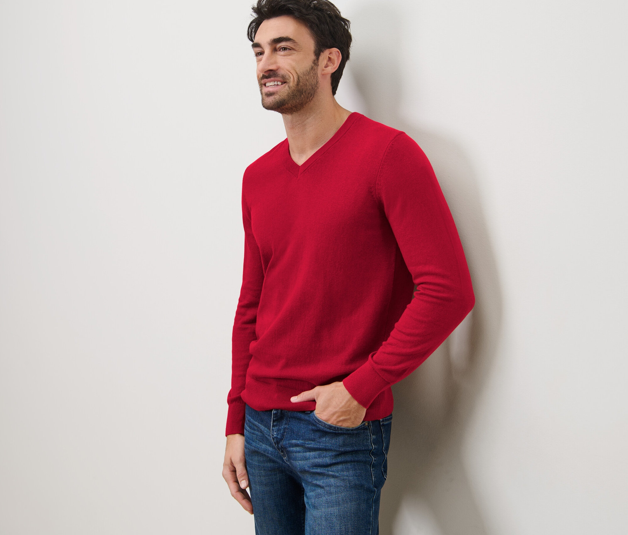 Duvara yaslanmış, kırmızı V yakalı kazak ve kot pantolon giyen gülümseyen bir adam.