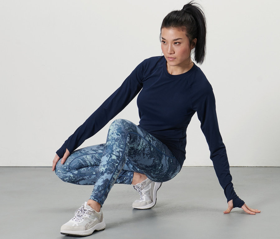 Koyu renkli üst ve Spor Taytı, koyu mavi/su yeşili giyen kadın çömelerek poz veriyor.