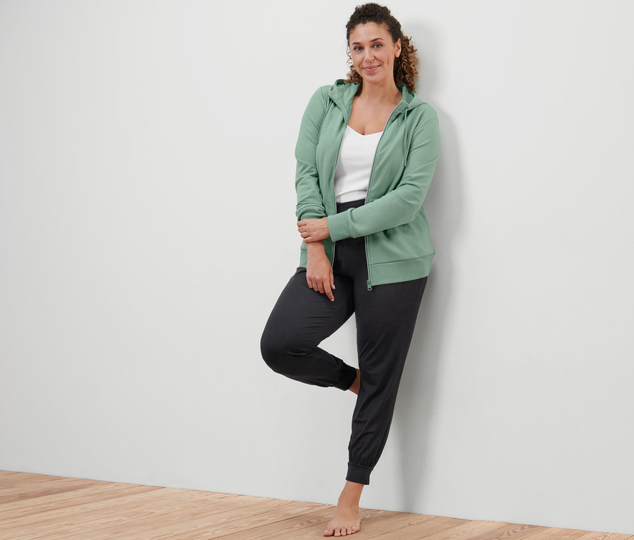 Bir kadın duvara yaslanmış, ada çayı yeşili renginde bir sweatshirt ceket ve yoga pantolonu giyiyor.