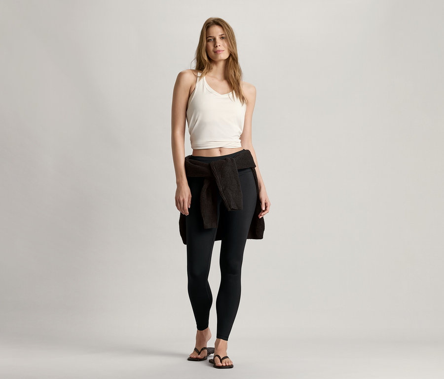 Kadın ayakta duruyor ve beyaz dikişsiz spor büstiyer, siyah tayt ve beline bağlanmış bir hırka giyiyor.