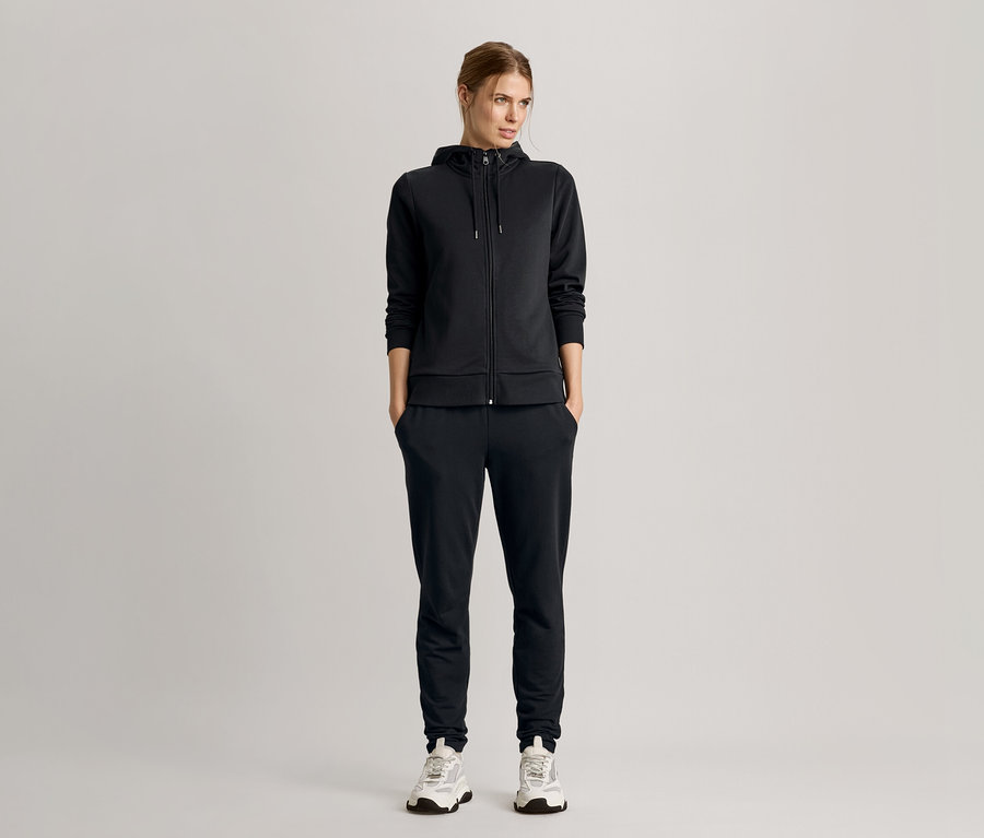 Kadın elleri cebinde duruyor, kapüşonlu ceket ve spor ayakkabılarla siyah bir eşofman takımı giyiyor.