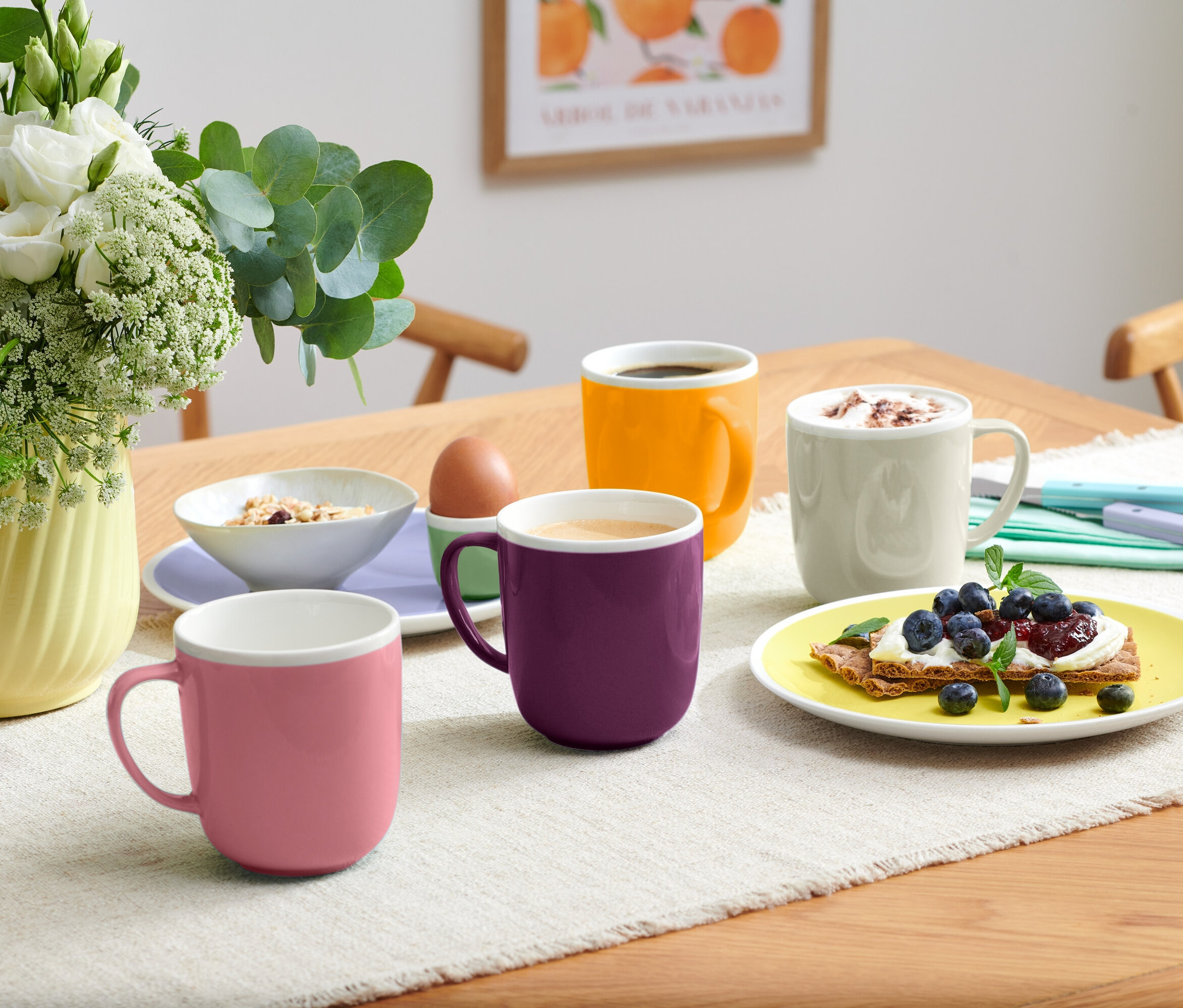 Bir masanın üzerinde dört farklı renkte kupa, bir kase müsli, bir yumurta ve yaban mersinli kızarmış ekmek bulunan bir tabak var.