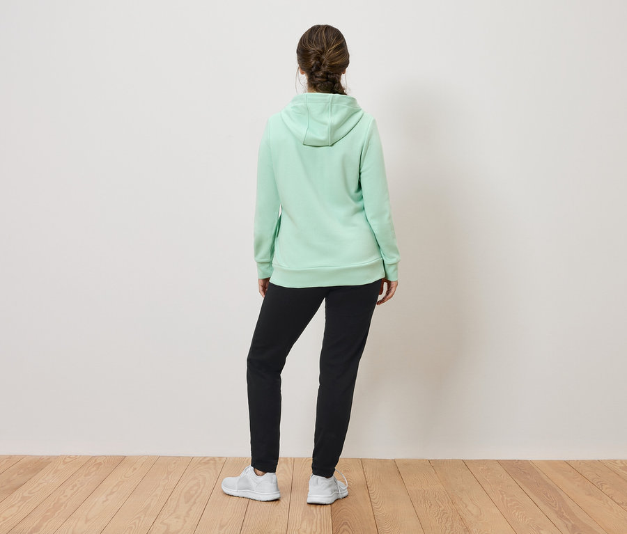 Sırtı kameraya dönük bir kadın, açık yeşil kapüşonlu bir sweatshirt, siyah eşofman altı ve beyaz spor ayakkabılar giyiyor.