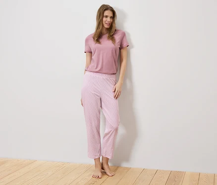 Bir kadın beyaz bir duvarın önünde duruyor, üzerinde pembe bir tişört ve desenli pembe bir pantolon var.