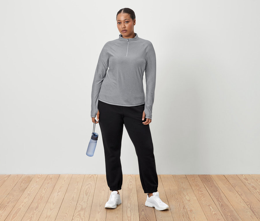 Bir kadın beyaz bir duvarın önünde duruyor ve elinde mavi bir su şişesi tutuyor. Gri uzun kollu bir tişört, siyah eşofman altı ve beyaz spor ayakkabı giyiyor.