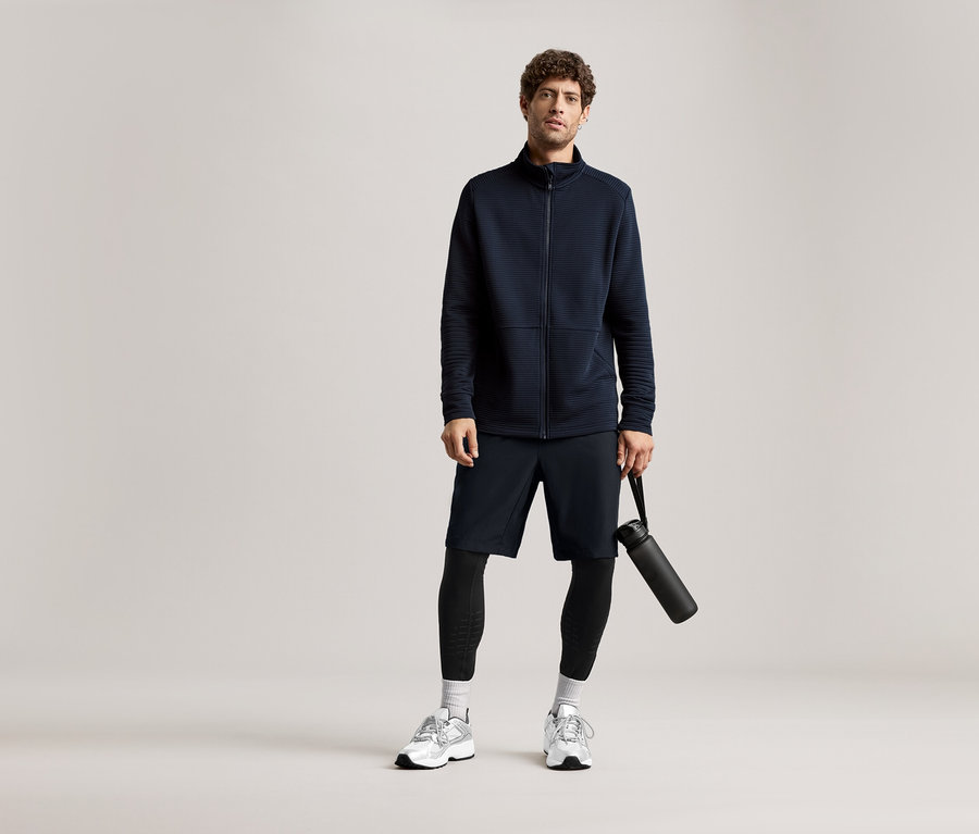 Lacivert ceket, şort ve tayt giyen bir adam siyah bir su şişesi tutuyor.