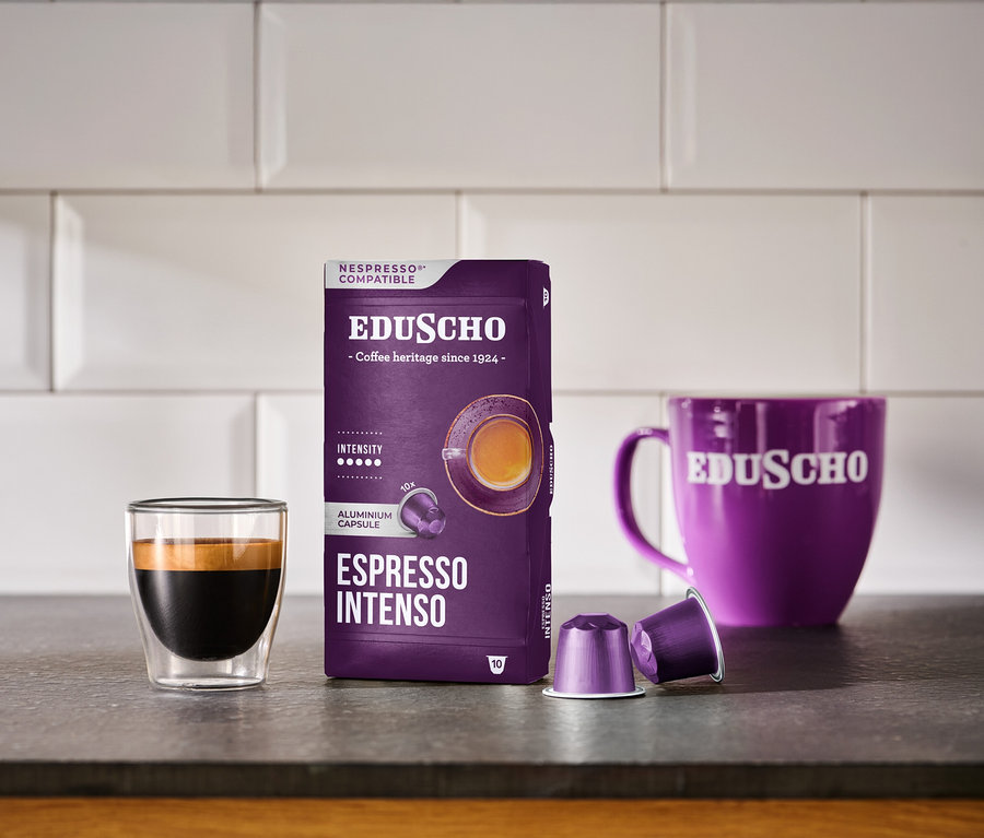 Mor renkli EDUSCHO Espresso Intenso Nespresso uyumlu kahve kapsülü kutusu, cam bir fincanda espresso, iki adet mor kapsül ve mor bir EDUSCHO kupası koyu renkli bir tezgah üzerinde beyaz fayansların önünde duruyor.