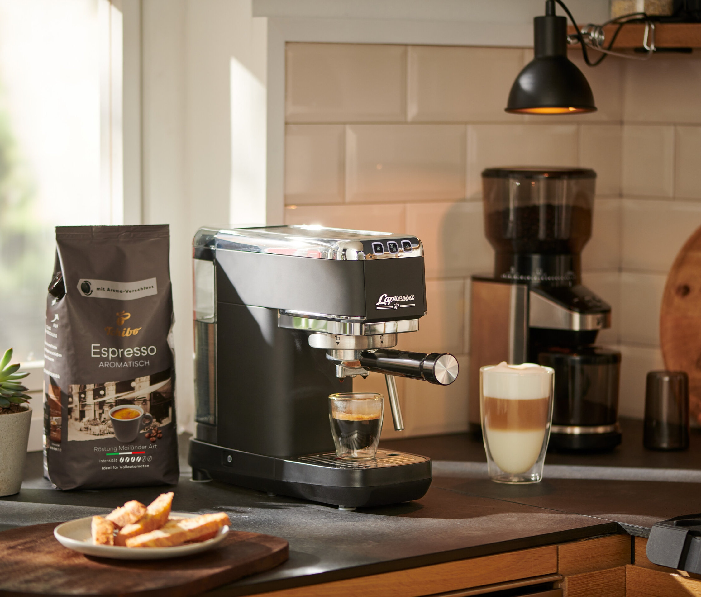 Espresso, siyah »Lapressa« Portafiltre Makinesi'nden bir bardağa akıyor. Arka planda bir kahve değirmeni ve bir paket kahve görülüyor.