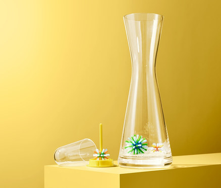 Şeffaf sürahi, sarı bir zemin üzerinde 3 adet şişe temizleme topu bulunan sarı bir tutucunun yanında duruyor.