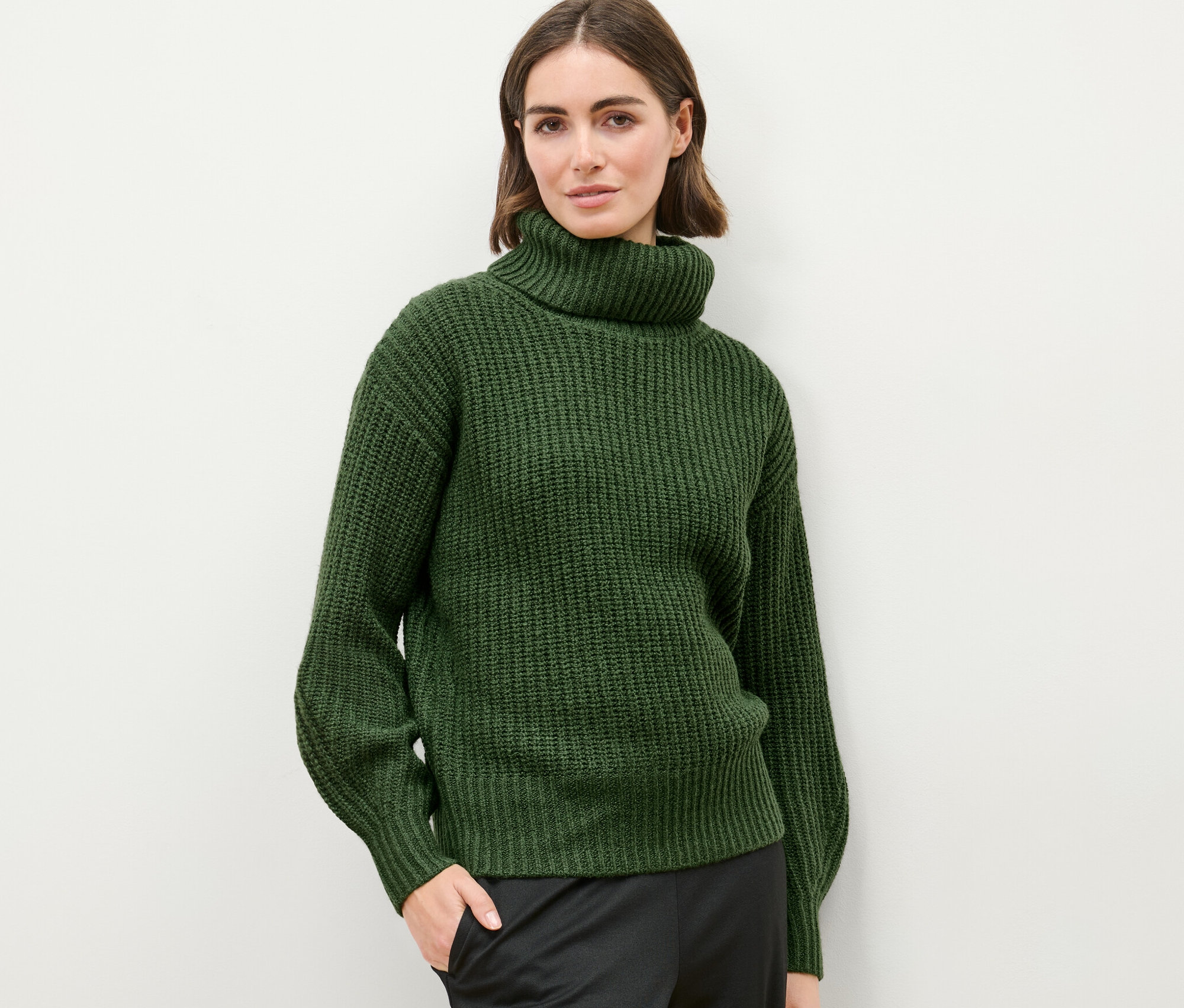 Açık renkli bir arka planda kalın örgülü boğazlı kazağıyla poz veren bir kadın.