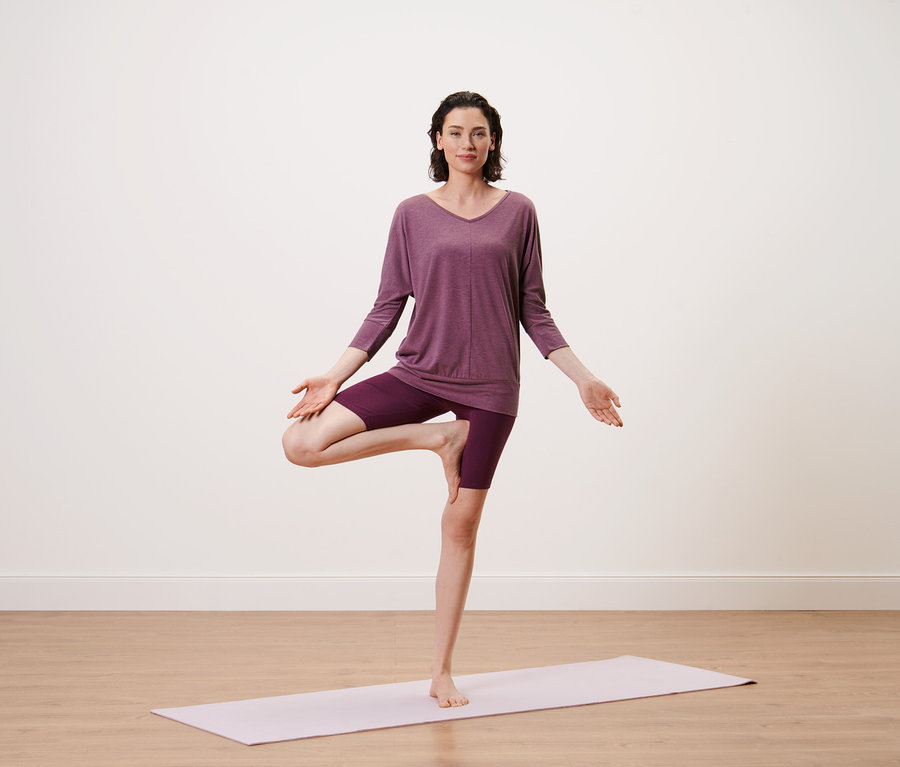 Yoga üstü, mürdüm rengi tişört giyen kadın yoga matı üzerinde ağaç pozisyonunda duruyor.