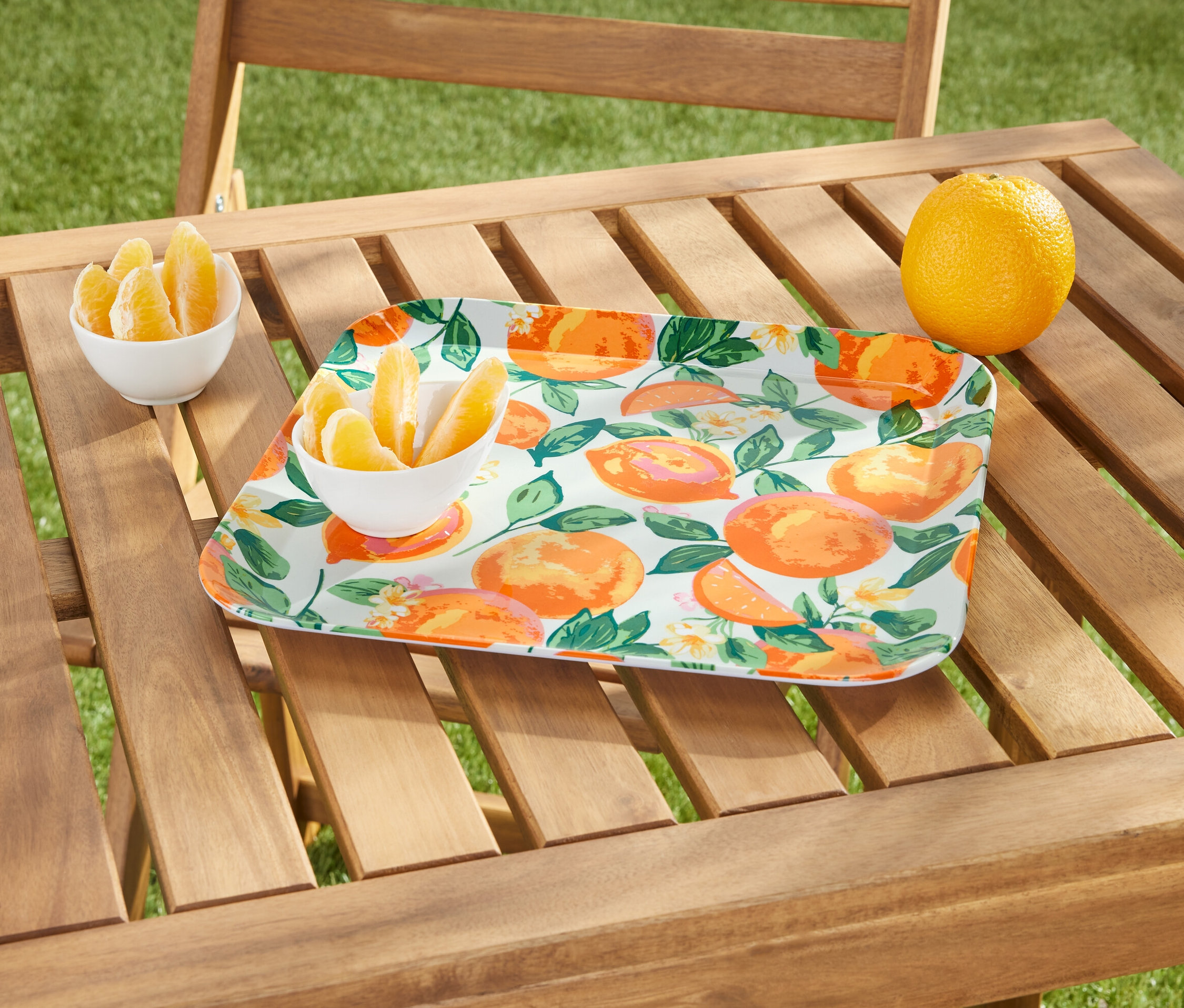 Ahşap bir masanın üzerinde portakal desenli bir servis tepsisi, bir kasede portakal dilimleri ve bütün bir portakal bulunmaktadır.