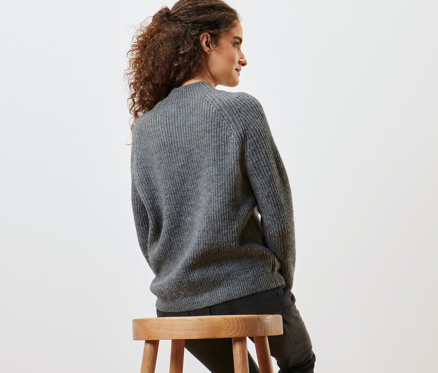 Bir kadın taburede oturuyor ve kırçıllı gri, dik yaka örgü kazak giyiyor.