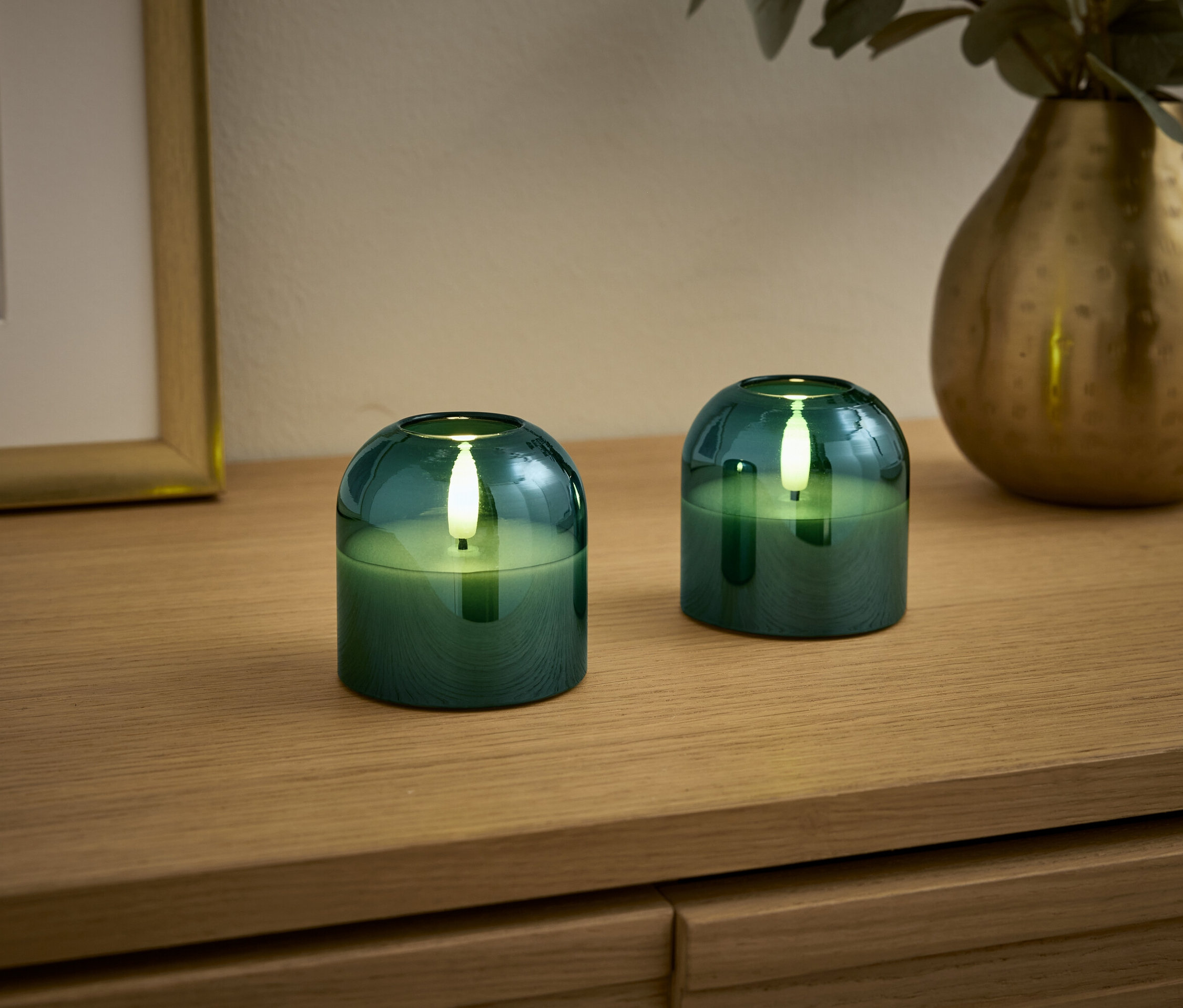 Ahşap bir yüzeyde iki adet yeşil mum, arka planda bir vazo ve bir resim çerçevesi bulunuyor.