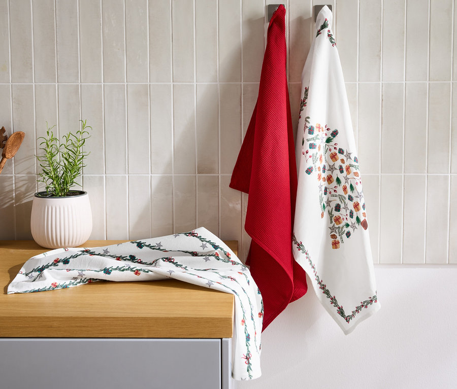 Duvara kırmızı bir bulaşık bezi ve Noel desenli beyaz bir bulaşık bezi asılı. Tezgahta Noel desenli beyaz bir bulaşık bezi var. Arka planda bitki saksısı ve tahta kaşıklar var.