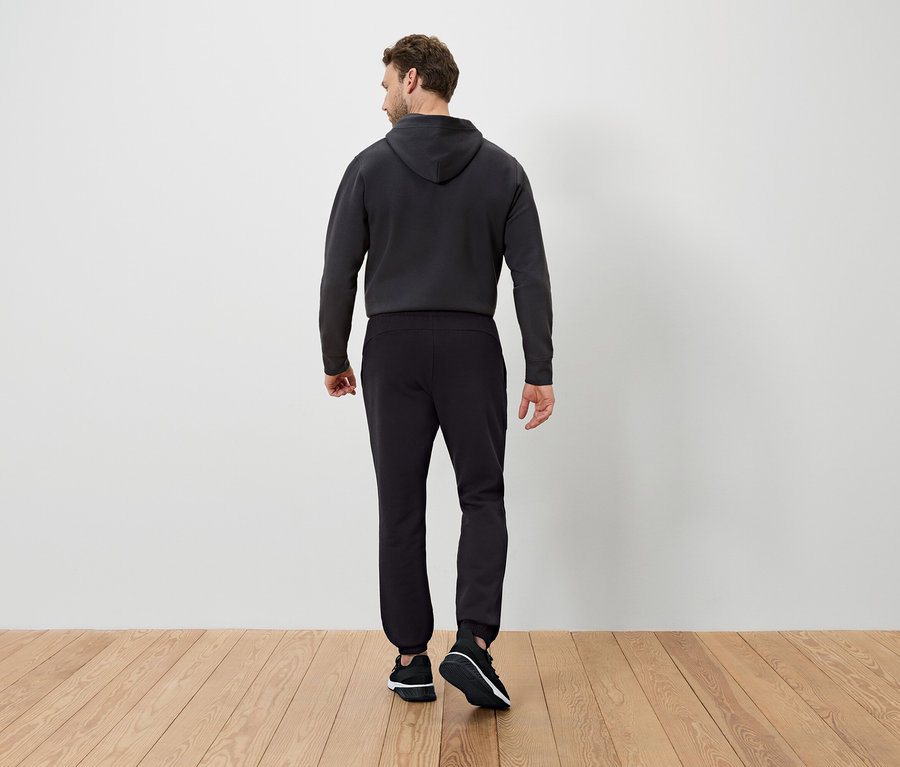 Arkası dönük bir adam koyu renk kapüşonlu bir sweatshirt ve siyah eşofman altı giyiyor.