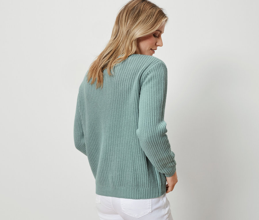 Arkası dönük bir kadın, adaçayı renginde, kalın örgülü bir hırka ve beyaz kot pantolon giyiyor.