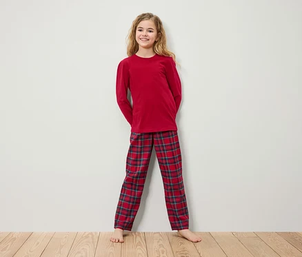 Kırmızı uzun kollu pijama ve ekose pantolonlu kız, beyaz bir duvarın önünde ahşap bir zeminde duruyor.