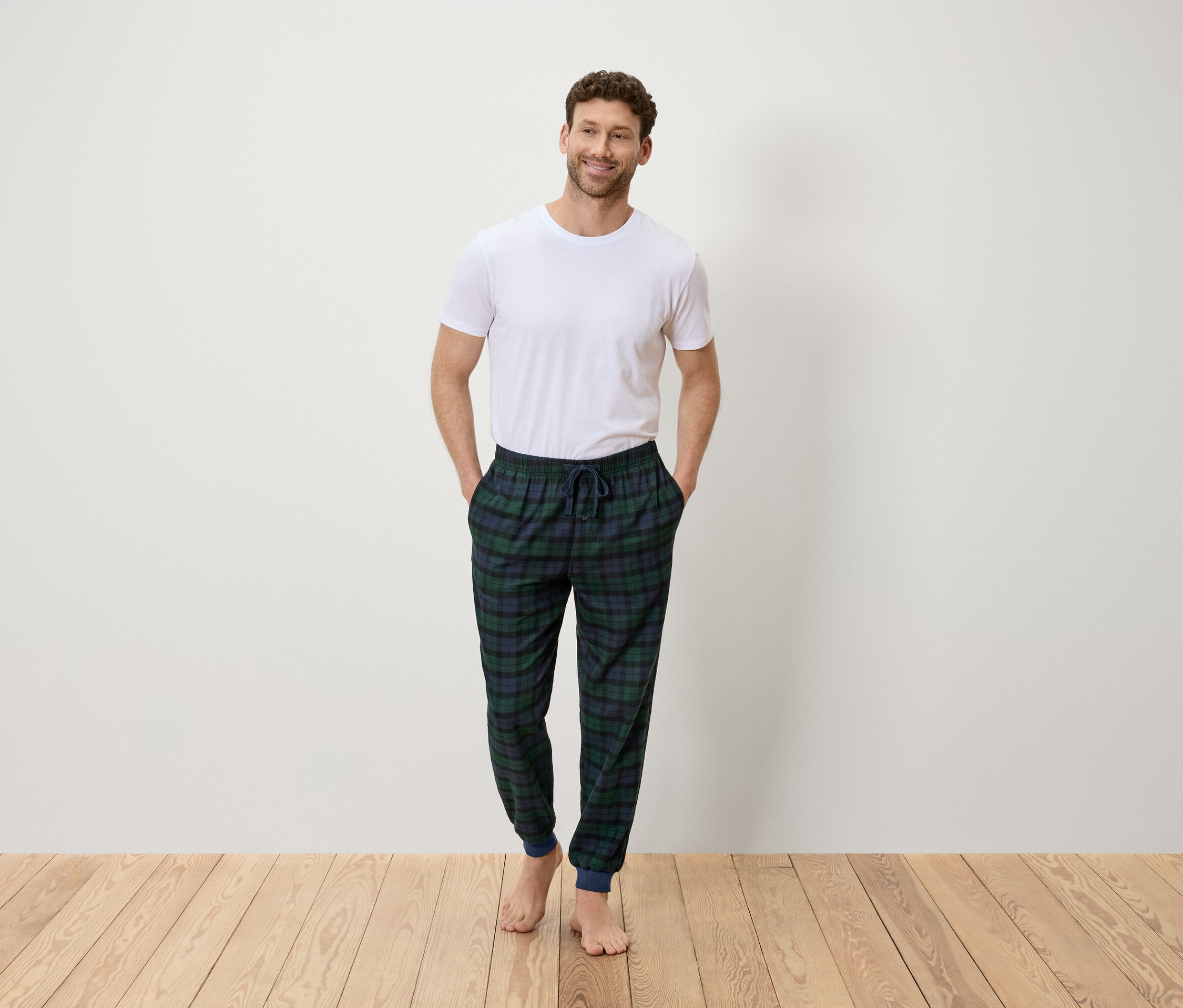 Adam beyaz tişört ve yeşil ve mavi ekose pantolon giymiş, elleri cebinde duruyor.
