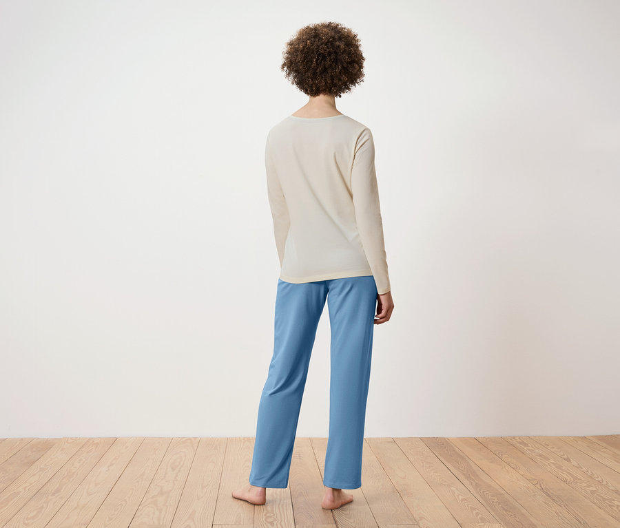 Arkadan, bej rengi uzun kollu bir tişört ve mavi pantolon giyen kıvırcık saçlı bir kadın görülüyor. Ahşap bir zeminde duruyor.