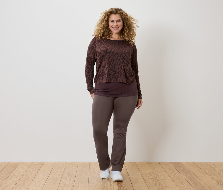 Kıvırcık saçlı bir kadın, kahverengi desenli uzun kollu bir bluz, daha koyu renk bir üst ve kahverengi pantolon ve beyaz spor ayakkabı giyiyor.