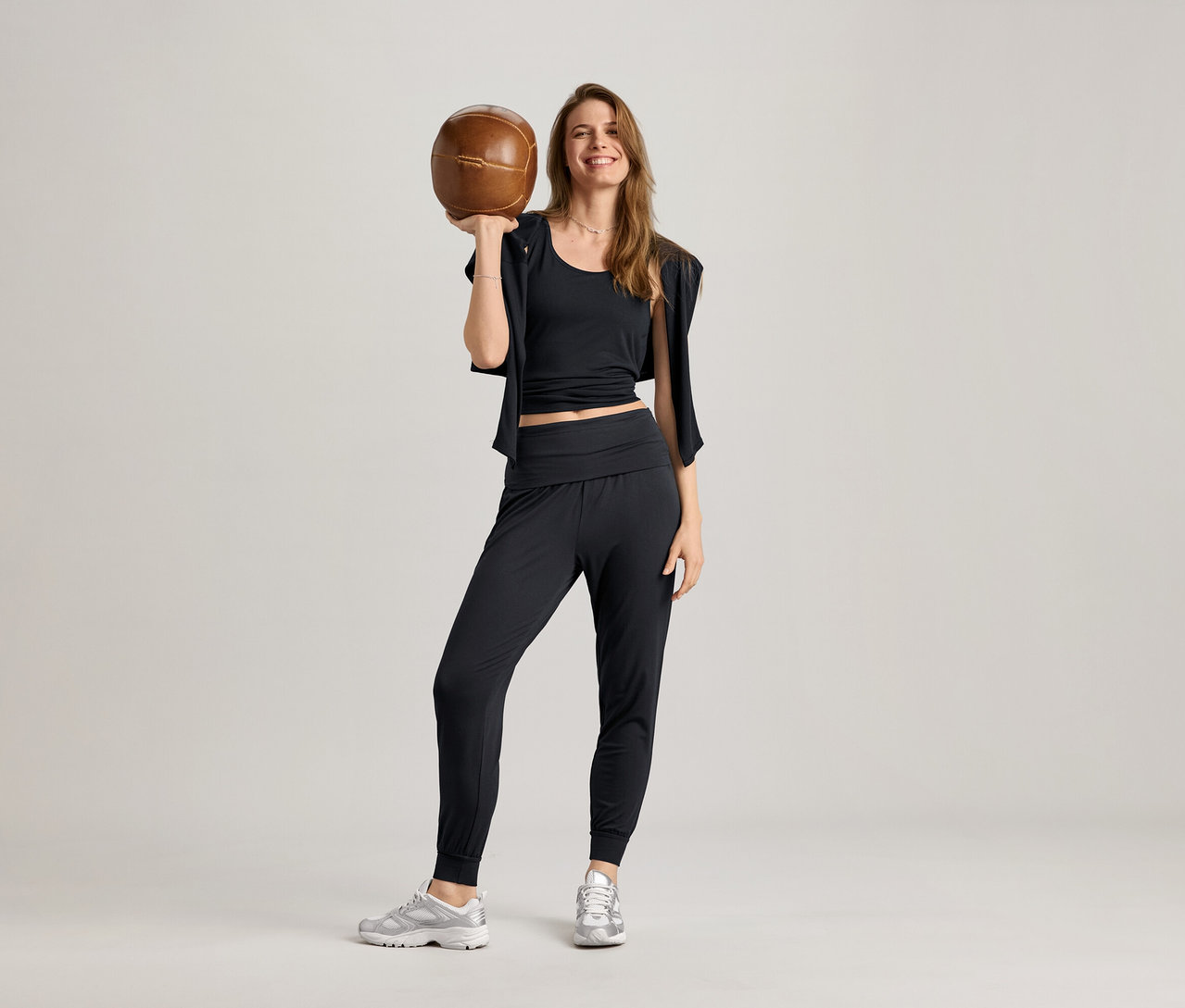 Siyah 2'si 1 arada spor tişörtü, eşofman altı ve spor ayakkabı giyen bir kadın kahverengi bir topu havaya kaldırıyor, gülümsüyor.