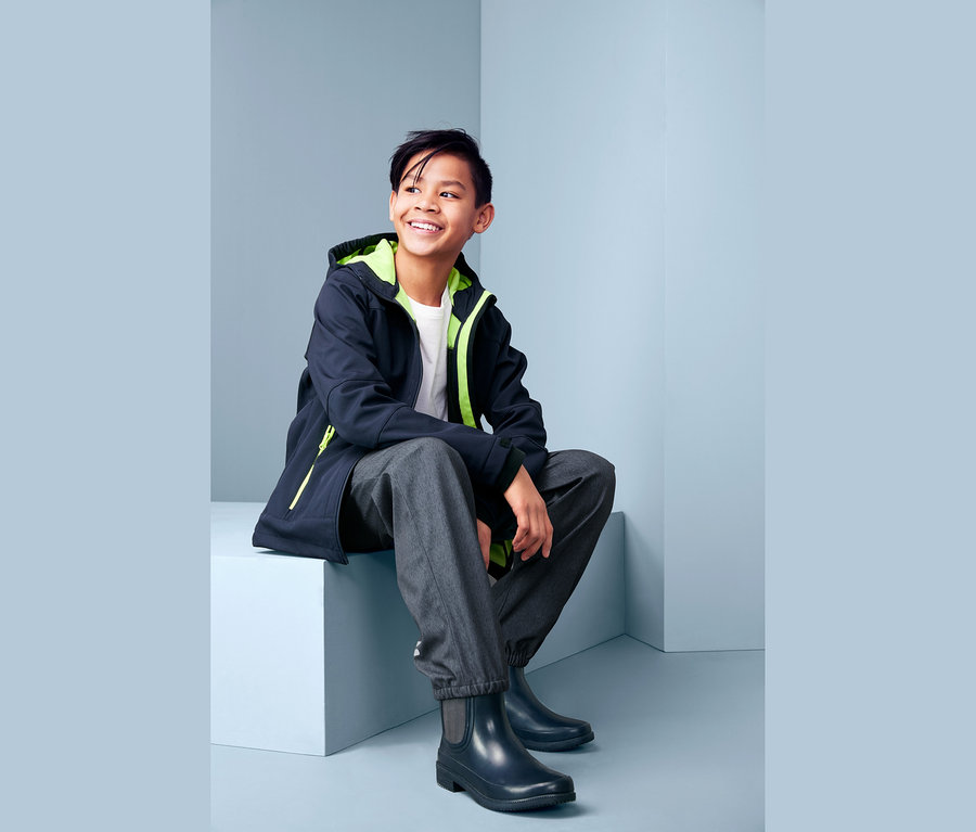 Gülümseyen bir çocuk oturuyor, üzerinde jean görünümlü softshell pantolon, ceket ve çizme var.