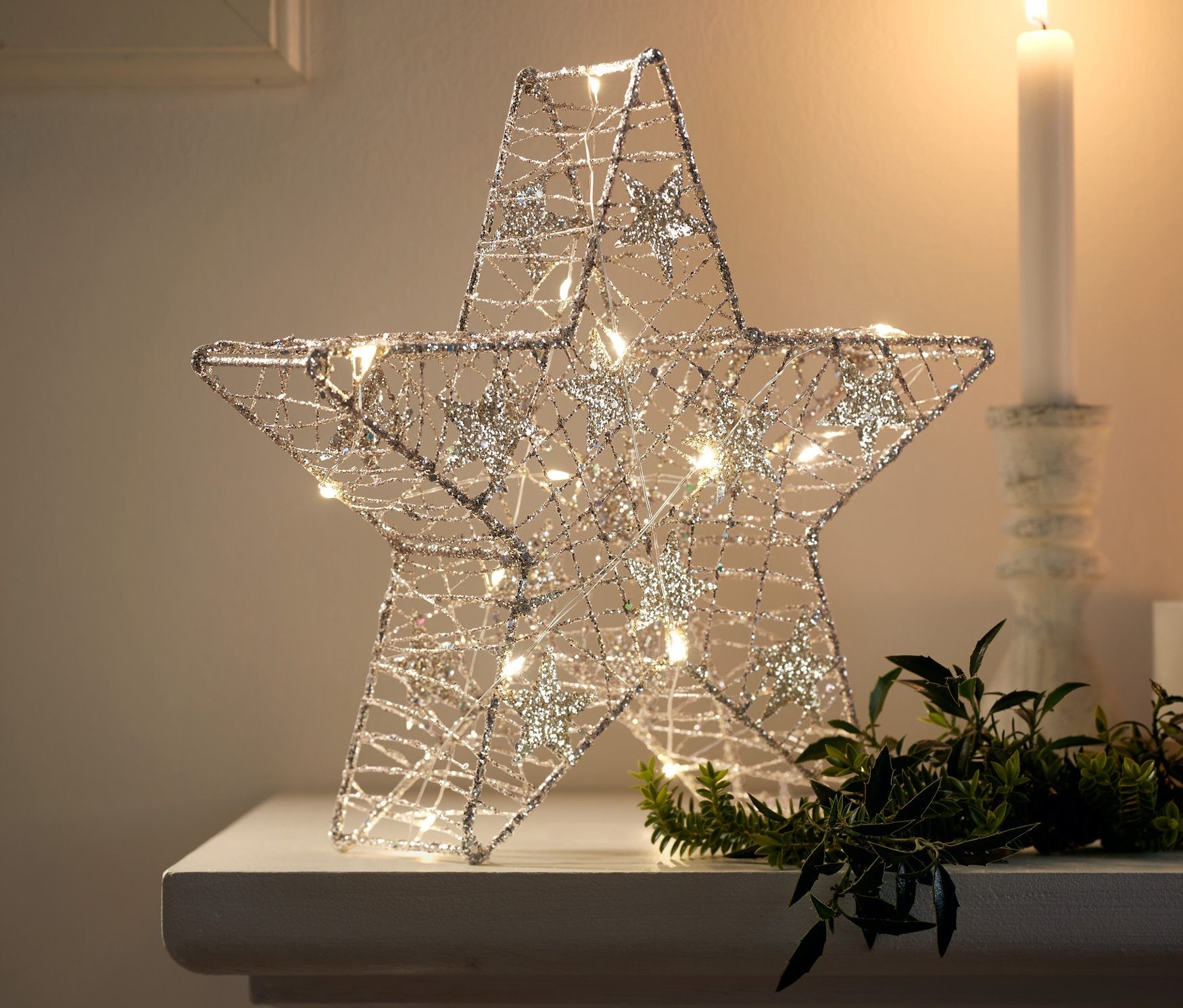 Altın rengi LED yıldız, beyaz bir yüzeyde yeşil bitkiler ve bir mumun yanında duruyor.