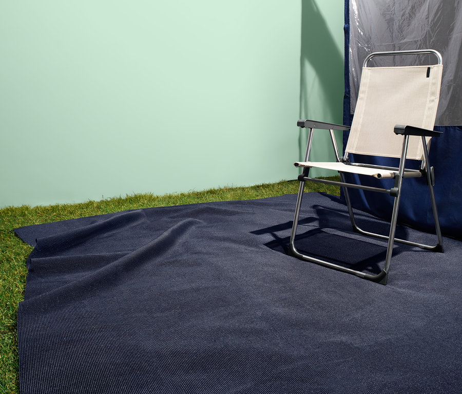 Koyu mavi çadır halısı, katlanır sandalye ve çadırın yanında çimenlerin üzerinde duruyor.